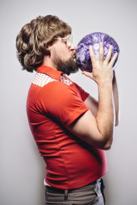 A shaggy haired man with a beard, glasses, and classic retro bowling shirt holds his ball, kissing it from a profile view. Vertical portrait; off white background with vignette.